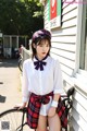 A woman in a white shirt and plaid skirt posing next to a bike.