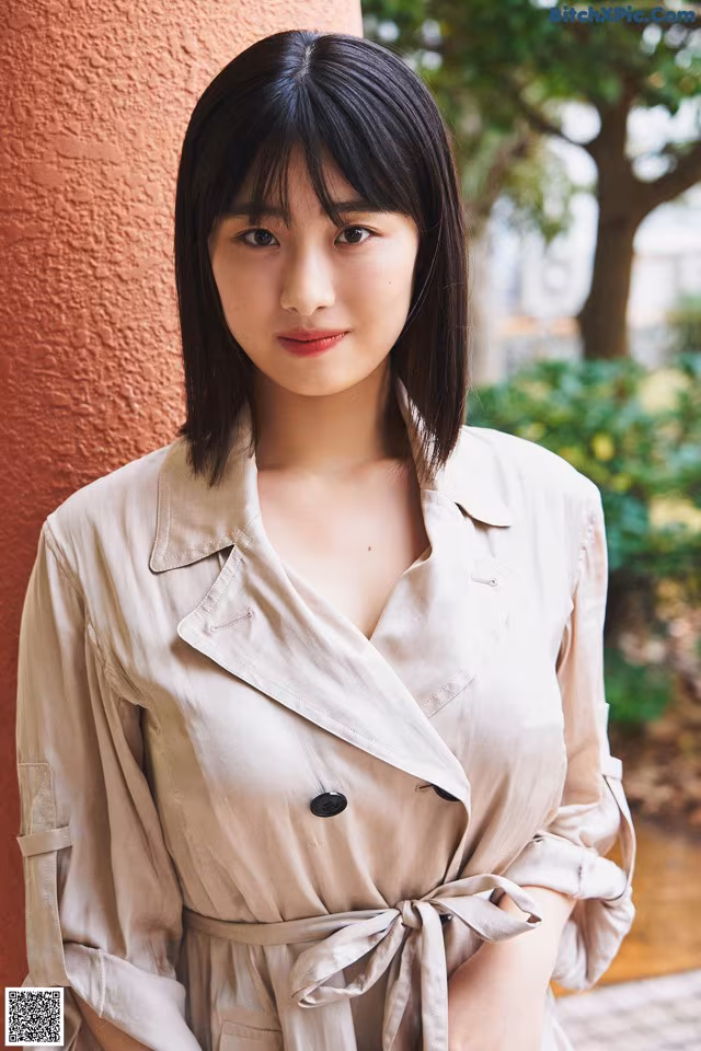A woman in a beige trench coat leaning against a wall.