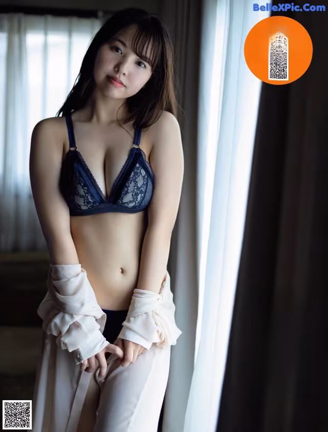 A woman in a blue bra and white pants posing by a window.
